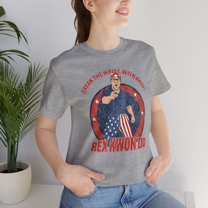 May include: Gray t-shirt featuring a cartoon illustration of a man in a blue shirt and American flag pants, pointing. Text reads "BREAK THE WRIST, WALK AWAY!" and "REX KWON DO".