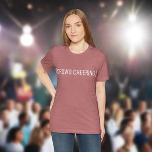 May include: A mauve-colored t-shirt with the text "[CROWD CHEERING]" in white. The shirt is being worn by a person in blue jeans. The background is blurred, suggesting a crowd of people.