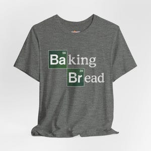 Könnte beinhalten: Graues T-Shirt mit dem Aufdruck "Baking Bread" im Periodensystem-Stil. "Baking" steht in einem grünen Quadrat mit der Nummer 56 darüber, und "Bread" steht in einem grünen Quadrat mit der Nummer 35 darüber. Das Design ist weiß.
