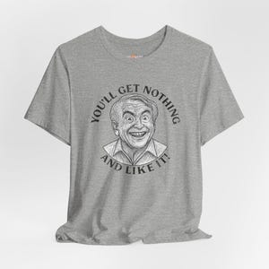 May include: Gray t-shirt featuring a black and white illustration of a smiling man with the text "YOU'LL GET NOTHING AND LIKE IT!" in a circular design around the image. The shirt has short sleeves and a crew neck.