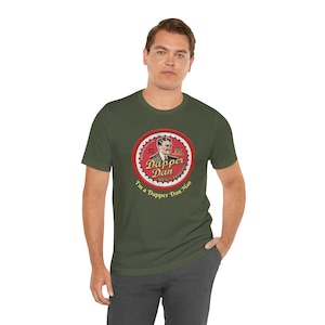May include: Olive green t-shirt featuring a circular graphic with the text "Dapper Dan Men's Pomade" and an illustration of a man. The text "I'm a Dapper Dan Man" is also included. The shirt is made of a soft material.