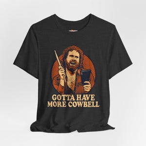 May include: Dark gray t-shirt featuring a retro-style illustration of a man holding a drumstick and a notepad. The image is set against a red circle, with the text "GOTTA HAVE MORE COWBELL" in a vintage font.