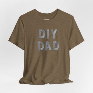 May include: A brown t-shirt with the words "DIY DAD" in silver duct tape style lettering. The shirt has short sleeves and a crew neck. The text is centered on the chest.