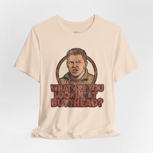 May include: Beige t-shirt featuring a distressed graphic of a man's face inside a red circle, with the text "WHAT ARE YOU LOOKIN AT BUTTHEAD?" in red and brown. The shirt has short sleeves and a crew neck.