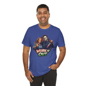May include: A blue t-shirt featuring a graphic of two people, one with a thumbs-up and the other holding a knife. The text "WORK IT OUT" is displayed in a colorful font. The shirt is made of a soft material.