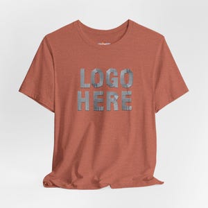May include: A rust-colored t-shirt with the words "LOGO HERE" in a metallic, textured font. The short-sleeved shirt is made of a soft material and is displayed on a neutral background.