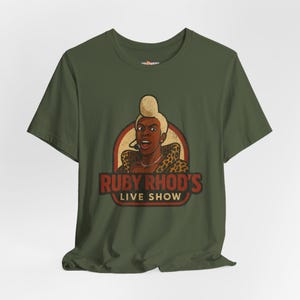 May include: Olive green t-shirt featuring a cartoon illustration of a person with a microphone, wearing a leopard print top. The text "RUBY RHOD'S LIVE SHOW" is displayed in a red and brown banner.