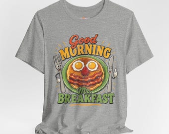 Puede incluir: Una camiseta gris con un diseño de desayuno de dibujos animados. El diseño incluye un plato de panqueques con tocino, huevos y una fresa, junto con las palabras "Good Morning Mr. Breakfast."