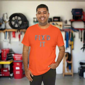 May include: An orange t-shirt with the text "FIX'D IT" in silver lettering. The short-sleeved shirt is made of a soft material. The person is wearing black pants.