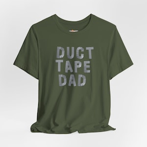May include: An olive green t-shirt with the text "DUCT TAPE DAD" in a silver, duct tape-like font. The shirt has a crew neck and short sleeves. The fabric is slightly wrinkled, suggesting it is made of cotton or a similar material.