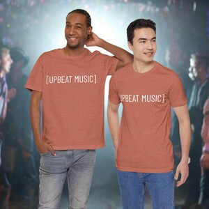 May include: Two people wearing rust-colored t-shirts with the text "[UPBEAT MUSIC]" in white. The shirts are short-sleeved and have a crew neck. The people are standing in front of a blurred background.