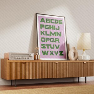 Puede incluir: Un gabinete de madera con un acabado de madera clara y un póster enmarcado de color rosa claro con el alfabeto en letras verdes sobre un fondo rosa claro. El póster dice "Don't Stop Believin". Hay una lámpara con una pantalla blanca y un jarrón de cerámica en la parte superior del gabinete.