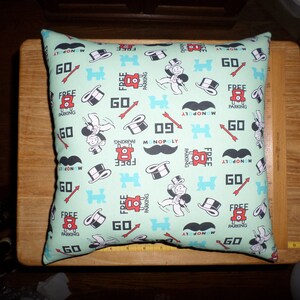 May include: Square pillow with a light green background featuring Monopoly game-themed graphics. The design includes images of the Monopoly man, top hats, mustaches, and the words "GO" and "FREE PARKING" in red and black.