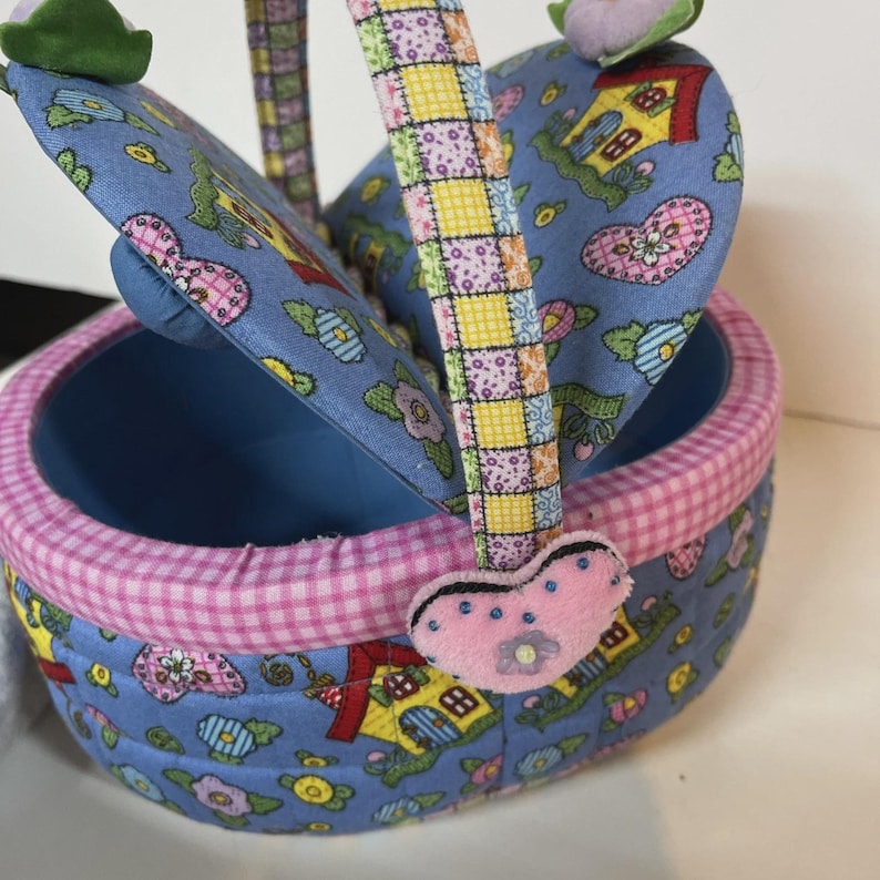 May include: A small, oval-shaped basket with a blue interior and a lid. The exterior features a blue fabric with a pattern of houses, flowers, and hearts. The rim is trimmed with pink gingham fabric, and the handle is patchwork. A pink heart-shaped charm hangs from the handle.