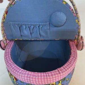May include: A blue fabric sewing basket with a floral and house pattern. The interior is blue, with a pink and white gingham trim. The lid has a pocket and a circular padded element. The handle and side accents are pink.