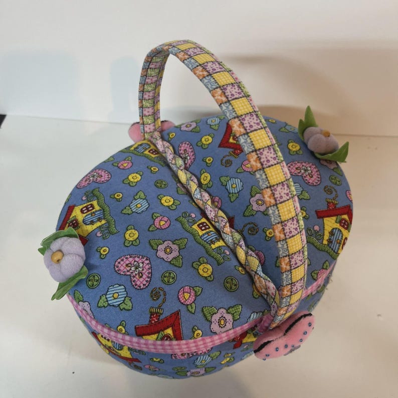 May include: A blue fabric basket with a handle, decorated with a pattern of houses, flowers, and hearts. The handle is yellow and pink checkered. The basket has pink and purple floral accents.