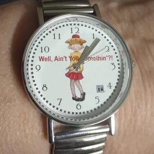 May include: A silver watch with a white face and black numbers. The watch face features a cartoon girl in a red dress and the text "Well, Ain't You Somethin'?!".