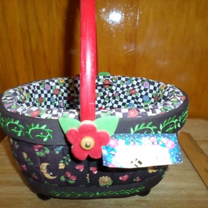 May include: A decorative basket with a red handle and a floral design. The basket has a black and white checkered interior and is adorned with a red flower and green leaves. The exterior features a dark brown fabric with embroidered green vines and colorful floral and heart motifs.