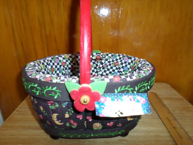 May include: A decorative basket with a red handle and a floral design. The basket has a black and white checkered interior and is adorned with a red flower and green leaves. The exterior features a dark brown fabric with embroidered green vines and colorful floral and heart motifs.