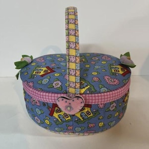 May include: A round, fabric-covered basket with a handle. The basket is primarily blue with a pattern of houses, flowers, and hearts. The handle is patchwork, and a pink heart-shaped embellishment is attached to the front. Two fabric flowers adorn the sides.