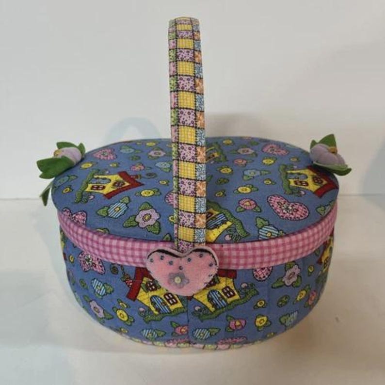 May include: A round, fabric-covered basket with a handle. The basket is primarily blue with a pattern of houses, flowers, and hearts. The handle is patchwork, and a pink heart-shaped embellishment is attached to the front. Two fabric flowers adorn the sides.