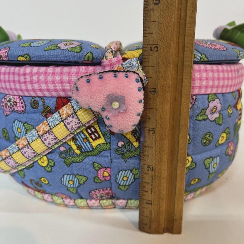May include: A round, quilted fabric basket with a blue floral pattern. The basket has a pink gingham trim and a pink heart-shaped embellishment. The basket is approximately 4 inches tall.