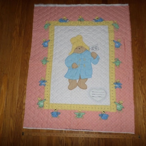 May include: A pink and white quilted fabric with a yellow border. The center features a cartoon image of a blue bunny wearing a yellow hat and holding a letter. The border is decorated with small blue bunnies.