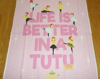 May include: Pink fabric with a white background featuring a girl in a yellow tutu and the text "Life is better in a tutu". The text is in pink and the girl is repeated in various poses around the text. The bottom of the fabric has the text "The Wiggles Emma!" in yellow.