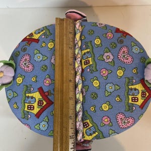 May include: A blue fabric decorative item with a house and heart pattern, accented with pink and yellow flowers. A wooden ruler is placed vertically, measuring approximately 6 inches. The item has a braided trim and pink accents.
