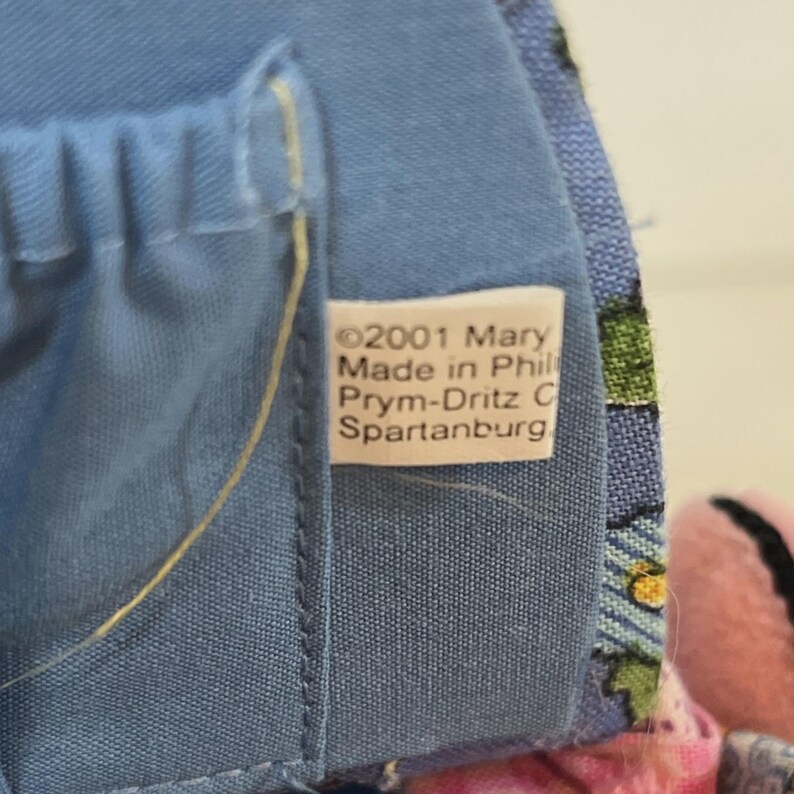 May include: Close-up of a blue fabric with a tag. The tag reads "&copy;2001 Mary Made in Phil Prym-Dritz C Spartanburg". The fabric is a solid blue color with a stitched seam. The tag is white with black text.