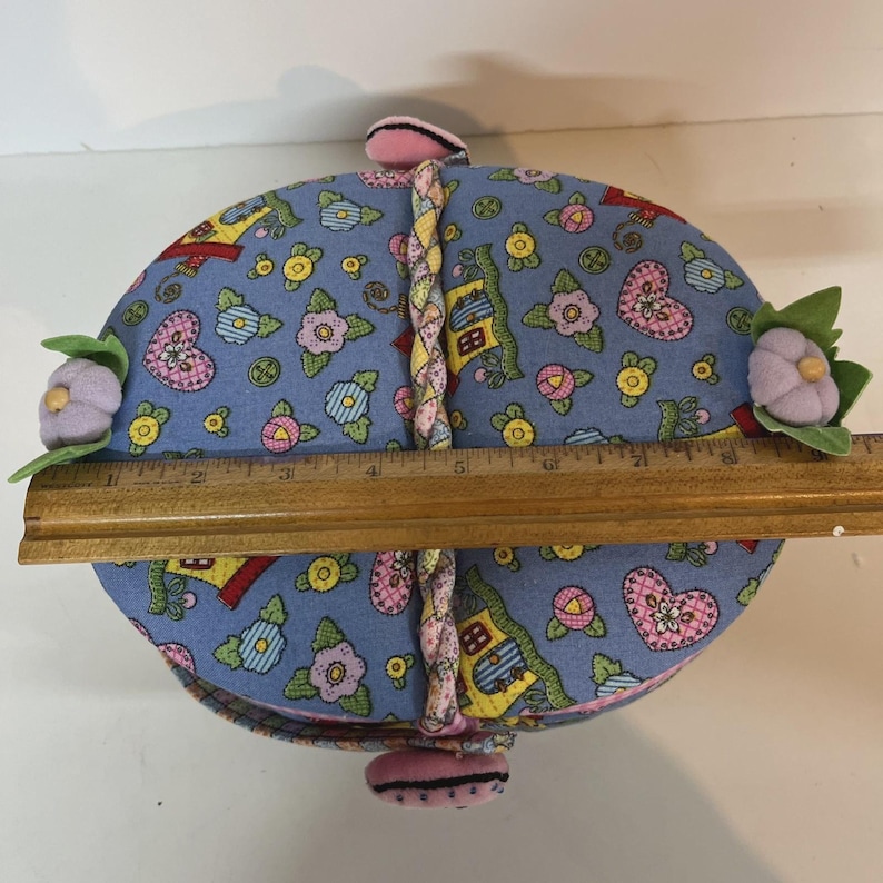 May include: A round, blue fabric container with a floral and heart pattern. The container is approximately 7 inches in diameter. It has a braided handle and is decorated with fabric flowers. The container is closed with a pink fabric closure.