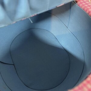 May include: An inside view of a blue paper container with curved edges. The container has a circular base and is partially open, revealing a pink and white patterned edge. The lighting creates shadows.