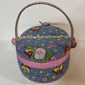 May include: A round, fabric basket with a handle, featuring a blue background with a pattern of houses and flowers. The basket has a pink gingham trim and is decorated with fabric flowers. The handle is multi-colored.