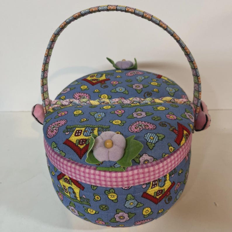 May include: A round, fabric basket with a handle, featuring a blue background with a pattern of houses and flowers. The basket has a pink gingham trim and is decorated with fabric flowers. The handle is multi-colored.
