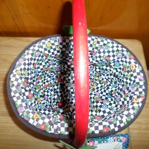 May include: An oval-shaped basket with a red handle. The interior features a floral pattern over a black and white checkered background. The basket's rim is trimmed with a brown border.
