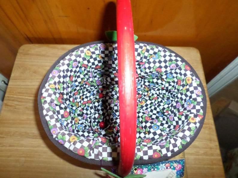 May include: An oval-shaped basket with a red handle. The interior features a floral pattern over a black and white checkered background. The basket's rim is trimmed with a brown border.