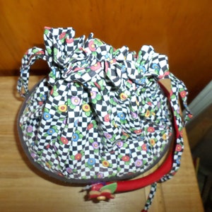 May include: A round, fabric handbag with a black and white checkered pattern, accented with colorful floral details. The bag has a red handle and a drawstring closure. The bag is approximately 8 inches in diameter.