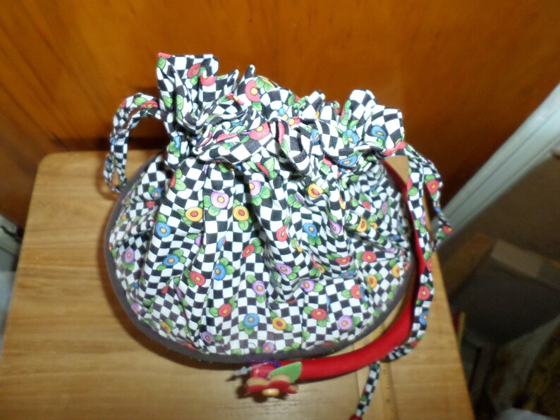 May include: A round, fabric handbag with a black and white checkered pattern, accented with colorful floral details. The bag has a red handle and a drawstring closure. The bag is approximately 8 inches in diameter.