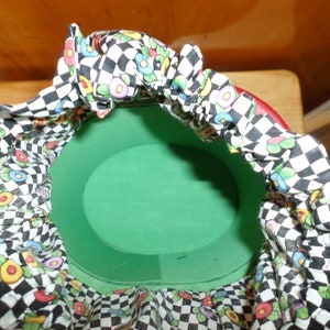 May include: A round, green container with a red rim, lined with fabric. The fabric features a black and white checkerboard pattern interspersed with colorful floral designs. The container is open, revealing its interior.