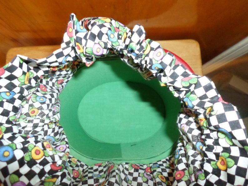 May include: A round, green container with a red rim, lined with fabric. The fabric features a black and white checkerboard pattern interspersed with colorful floral designs. The container is open, revealing its interior.