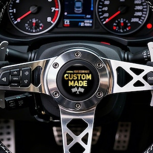 May include: Black suede steering wheel with white stitching and a silver metal center. The center features the text "CUSTOM MADE" in a black circle. The car's dashboard and gauges are visible in the background.