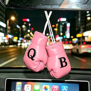 May include: A pair of miniature pink boxing gloves hanging from a rearview mirror. Each glove features a single black letter printed on the front, with the left glove displaying the letter Q and the right glove displaying the letter B.