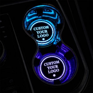 May include: A pair of circular car cup holder inserts featuring bright blue LED illumination. Each insert displays the text CUSTOM YOUR LOGO in white lettering centered within a glowing circular border, set against a dark interior console.