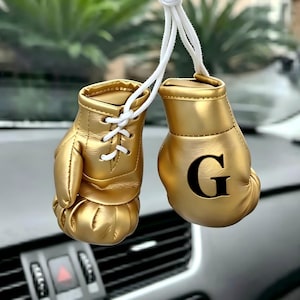 May include: A pair of miniature gold boxing gloves hanging from white cords. One glove features a black letter G printed on the front, while the other glove has white laces. The gloves have a smooth, metallic finish and are designed as a hanging ornament.
