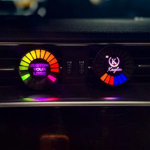 Puede incluir: Dos difusores de aroma circulares para rejillas de ventilación de coche con iluminación LED integrada. La unidad izquierda muestra el texto CUSTOM YOUR LOGO en morado, mientras que la derecha muestra una letra K blanca en una corona.