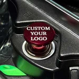May include: A circular, metallic dark red car cigarette lighter plug featuring the white text CUSTOM YOUR LOGO printed on the top surface. The plug is inserted into a black plastic vehicle console socket with a metallic silver rim.