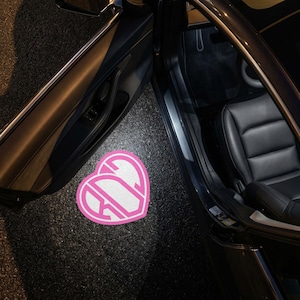 May include: A pink and white heart-shaped car floor mat with a monogram design. The mat is placed on the asphalt next to a car door. The car's interior features black leather seats.