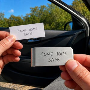 Op de afbeelding: Een rechthoekige geldclip van geborsteld metaal met de gegraveerde tekst COME HOME SAFE in een eenvoudig schreefloos lettertype. De clip wordt vastgehouden naast een kleine witte strook papier die ook de handgeschreven tekst COME HOME SAFE toont.