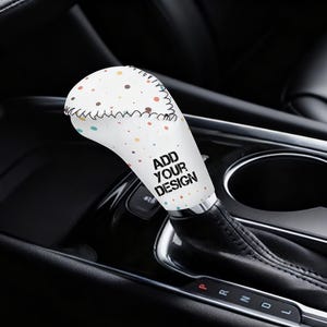 Könnte beinhalten: Ein weißer Schalthebelbezug mit einem Muster aus kleinen, farbigen Punkten. Der Bezug trägt den Text "ADD YOUR DESIGN" in Schwarz. Der Schalthebel befindet sich in einem Autoinnenraum mit einem schwarzen Lederschalthebel.