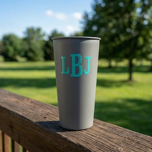 May include: A gray tumbler with the initials "LBJ" in teal. The tumbler is cylindrical and appears to be made of metal. It is sitting on a wooden surface with a green background.